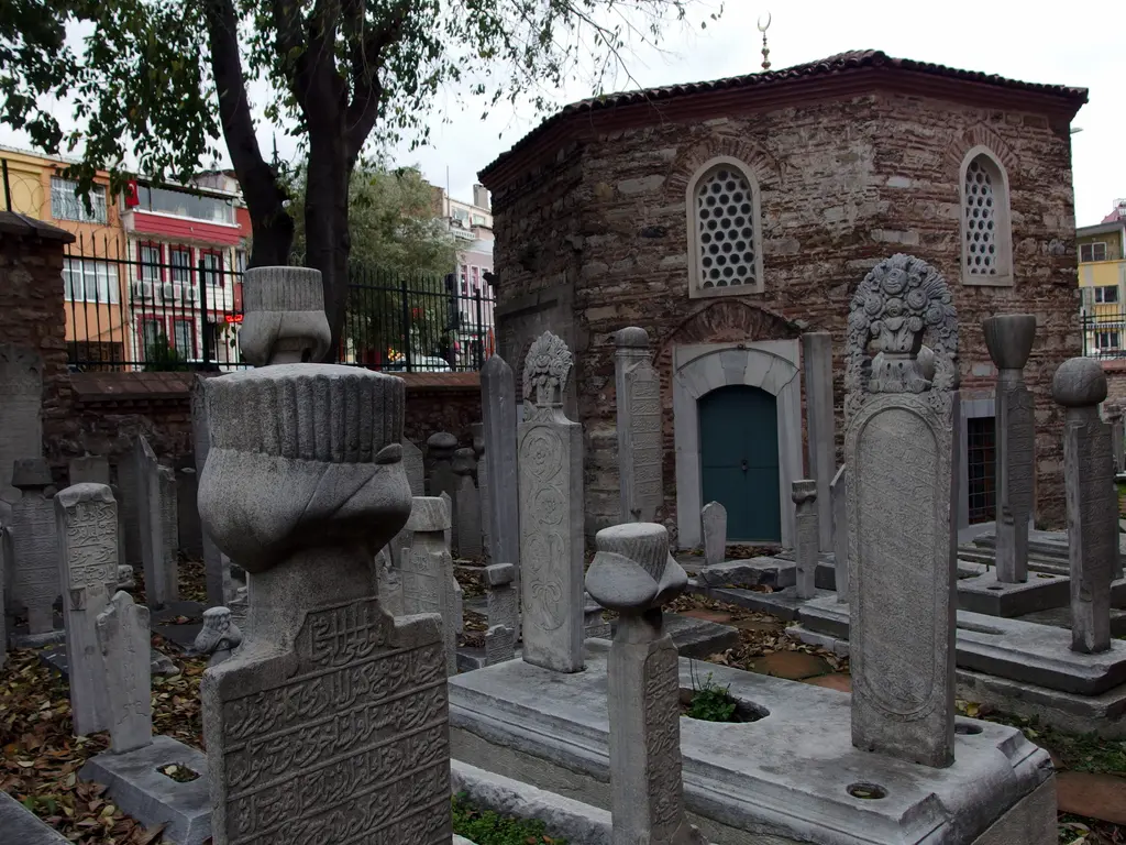 Hazirede Hüseyin Ağa'nın türbesi; önde kavuk başlıklı gri mezar taşları, arkada sekizgen planlı, tuğla taş almaşık duvarlı, sivri kemerli pencereli ve yeşil kapılı kubbeli türbe.