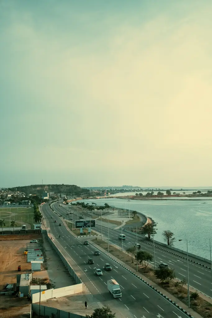 Luanda'da körfez boyunca kıvrılan geniş sahil otoyolu; palmiyeli orta refüj, deniz kıyısı ve puslu sıcak ışıkta ilerleyen araçlar, yüksekten bakış.