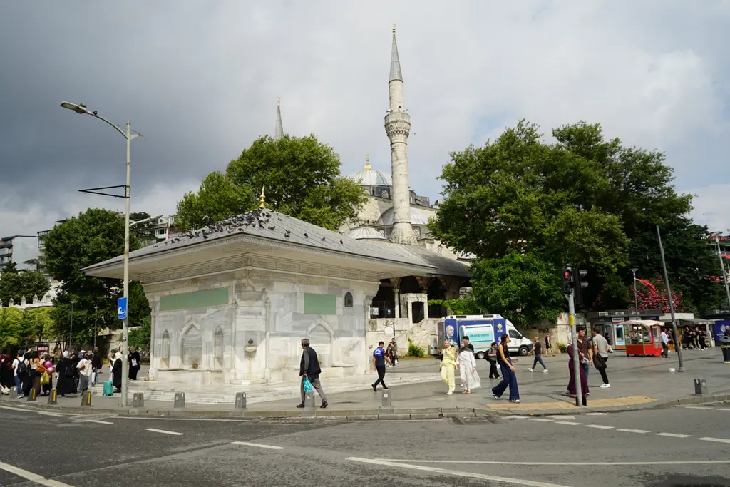 Üsküdar'da III. Ahmed Çeşmesi; meydanda mermer çeşme, arkada ağaçlar, cami minaresi ve yürüyen insanlar.