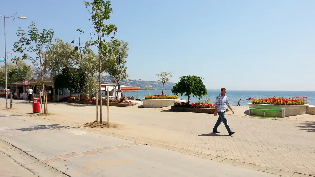 Büyükçekmece sahil yolu; taş döşeli meydan, banklar, çiçek saksıları, yürüyen bir adam ve arkada deniz.