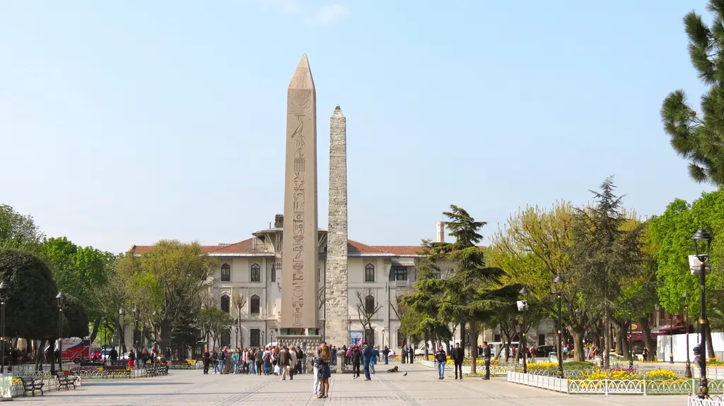 Sultanahmet Meydanı'nda Dikilitaş ile arkasında Örme Sütun; önde kalabalık, çiçek tarhları, arkada İbrahim Paşa Sarayı'nın cephesi.