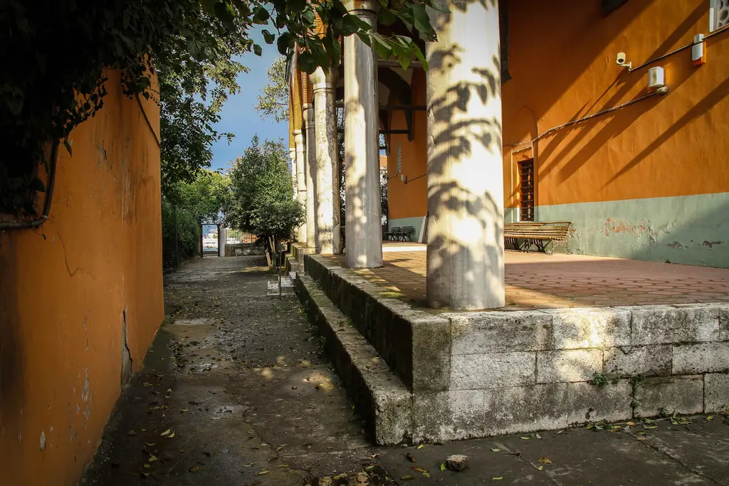 Rum Mehmed Paşa Camii'nin yan cephesi; turuncu duvar, revak sütunları, taş kaide ve yanından geçen dar yol.