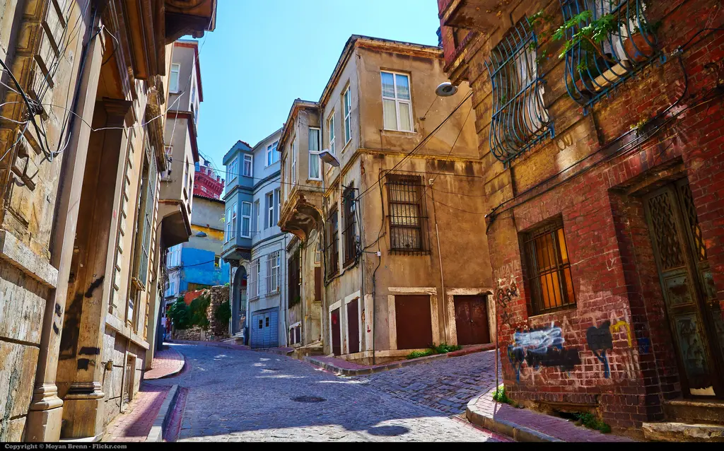 Balat'ta arnavut kaldırımlı dar sokak; iki yanda eski taş ve ahşap evler, cumbalar ve mavi gökyüzü.