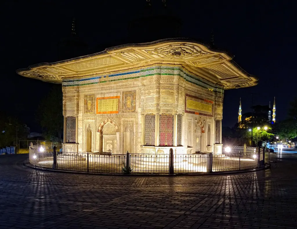 Gece aydınlatılmış çeşme; sarı ışıkla parlayan mermer cepheler, karanlıkta seçilen saçak ve uzakta aydınlatılmış minareler.