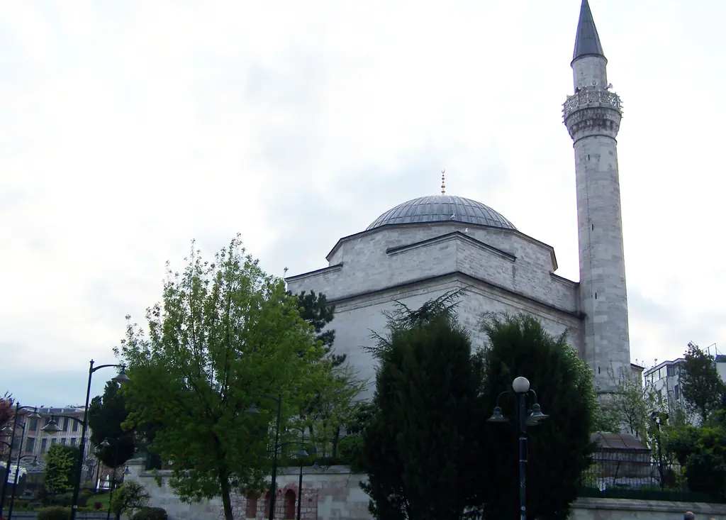 Firuz Ağa Camii'nin kurşun kubbesi ve taş minaresi; önde yeşil ağaçlar, bir sokak lambası ve bulutlu açık gökyüzü.