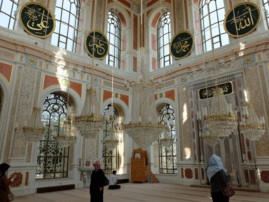 Ortaköy Camii'nin harimi; ortada mihrap, iki yanda büyük pencereler, avizeler, yeşil hat levhaları ve birkaç ziyaretçi.