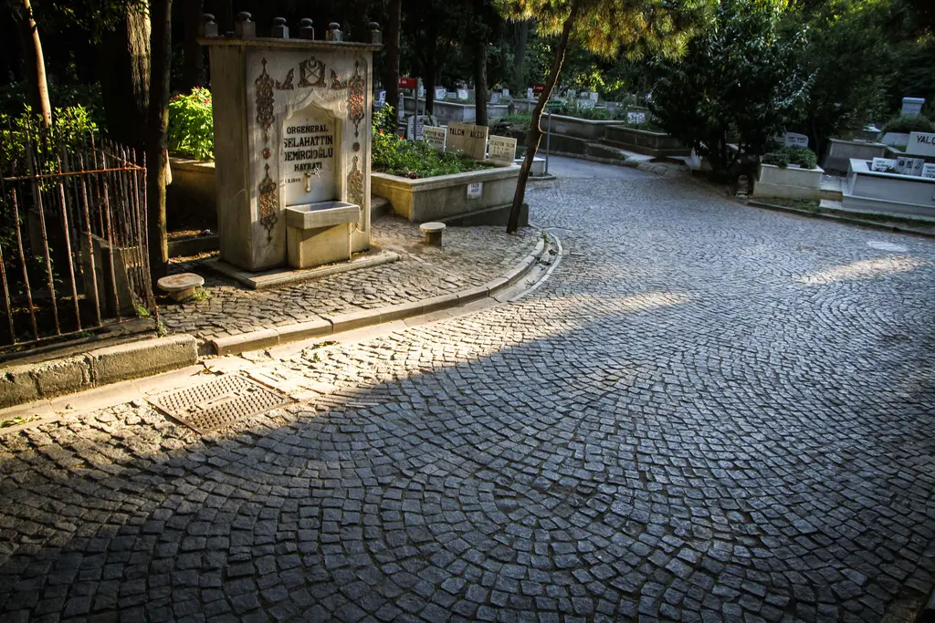 Karacaahmet'te çeşme; kitabeli taş ayna, yanında demir parmaklık, önünde parke taşı yol ve arkada kabirler.