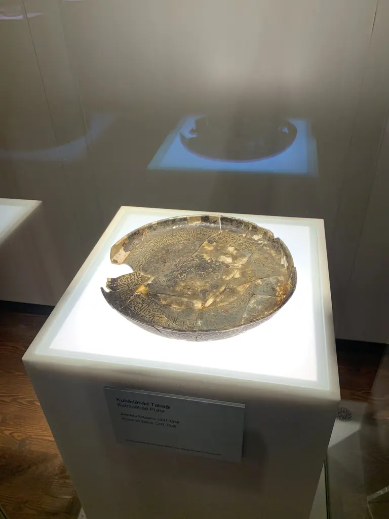 Beykoz Cam ve Billur Müzesi'nde ışıklı bir kaide üzerinde, kenarı kırık ve yüzeyi altın yaldız kalıntılı yuvarlak bir cam tabak.