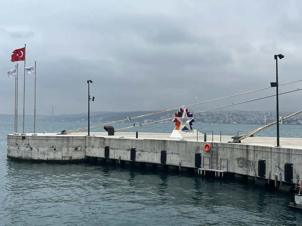 Galataport'ta denize uzanan beton iskele; bayrak direkleri, aydınlatma direkleri ve uzakta Boğaz Köprüsü.