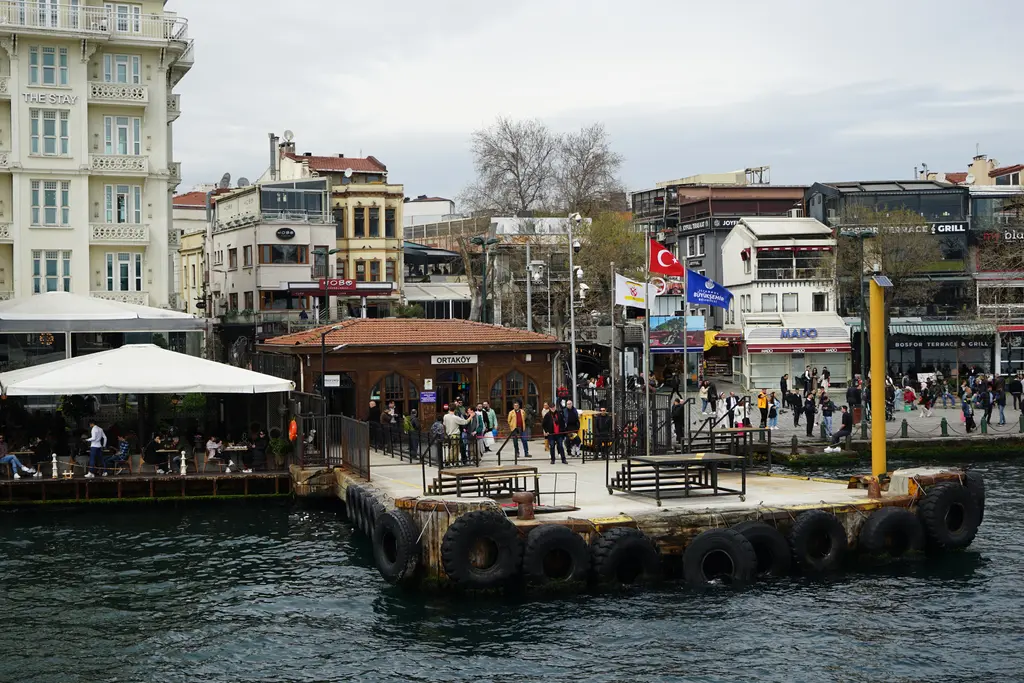 Ortaköy iskelesi; önde iskele ve beyaz tenteli kafe, arkada renkli cepheli binalar, dalgalanan bayraklar ve yürüyen insanlar.