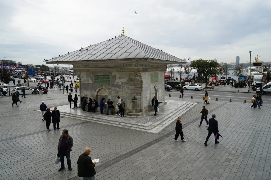 Üsküdar III. Ahmed Çeşmesi; taş döşeli meydan, çatıda güvercinler, çeşmenin çevresinde dolaşan insanlar.