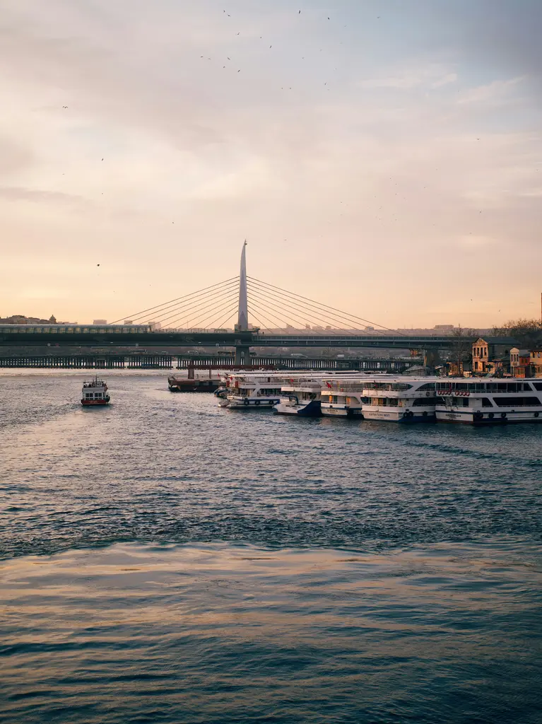 Haliç'ten Haliç Metro Köprüsü; ortadaki beyaz kuleye bağlı eğik halatlar, kıyıda bağlı gezi tekneleri ve akşam ışığında pembe gökyüzü.