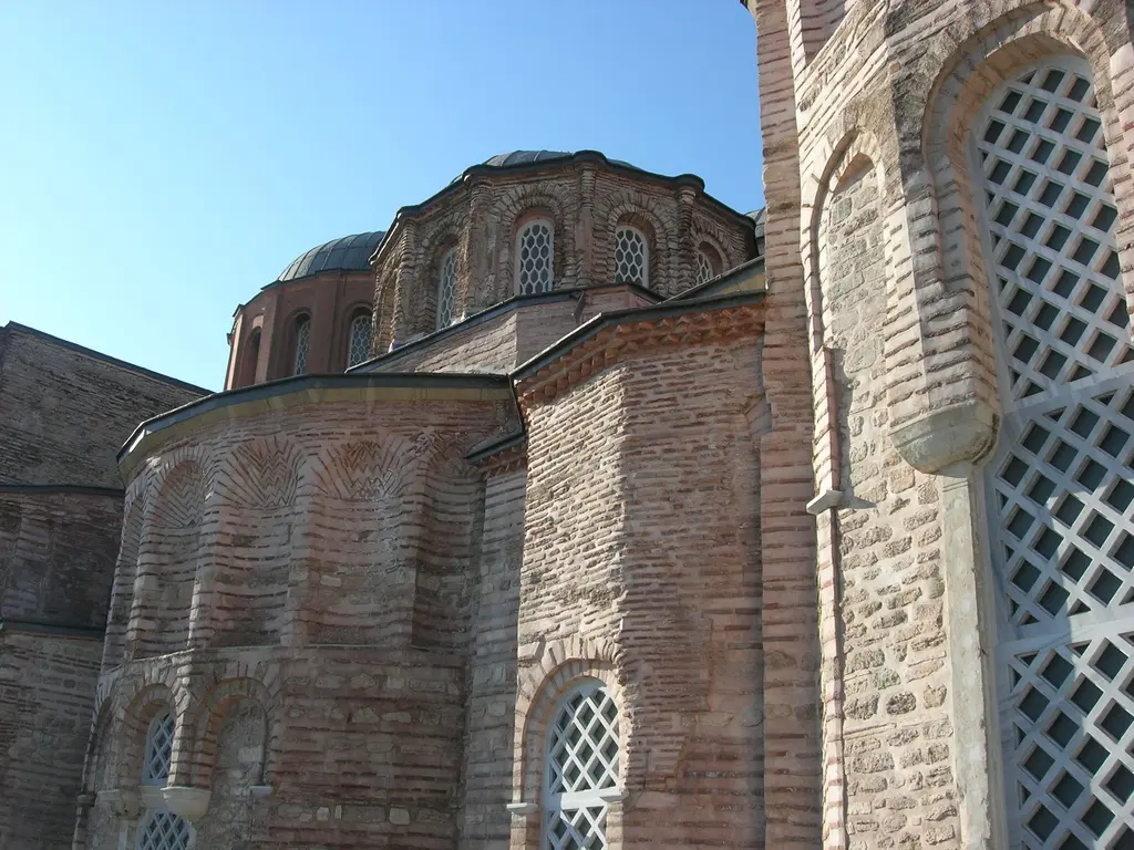 Zeyrek Camii'ne alttan bakış; önde apsisin tuğlayla örülmüş zikzak bezemesi, arkada kurşun kaplı iki kubbe, beyaz kafesli kemerli pencereler ve berrak mavi gökyüzü.