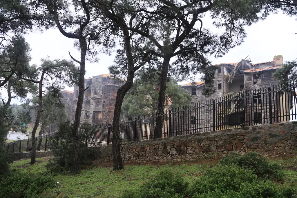 Büyükada Rum Yetimhanesi; taş duvarın ve ağaçların ardında ahşap kanatlar ve yeşil bahçe.