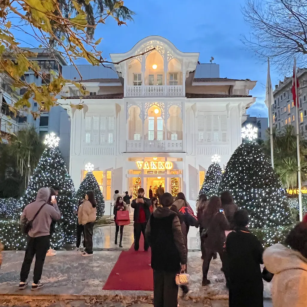 Aralıkta Vakko köşkü; beyaz ahşap köşk cephesi, ışıklı yılbaşı ağaçları, kırmızı halı ve fotoğraf çeken ziyaretçiler.