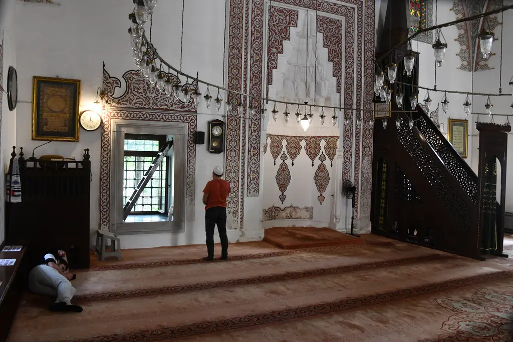 Firuz Ağa Camii'nin harimi; mihrap, ahşap minber, kalem işi süslü duvarlar, sarkan kandil halkası ve namaz kılan bir kişi.