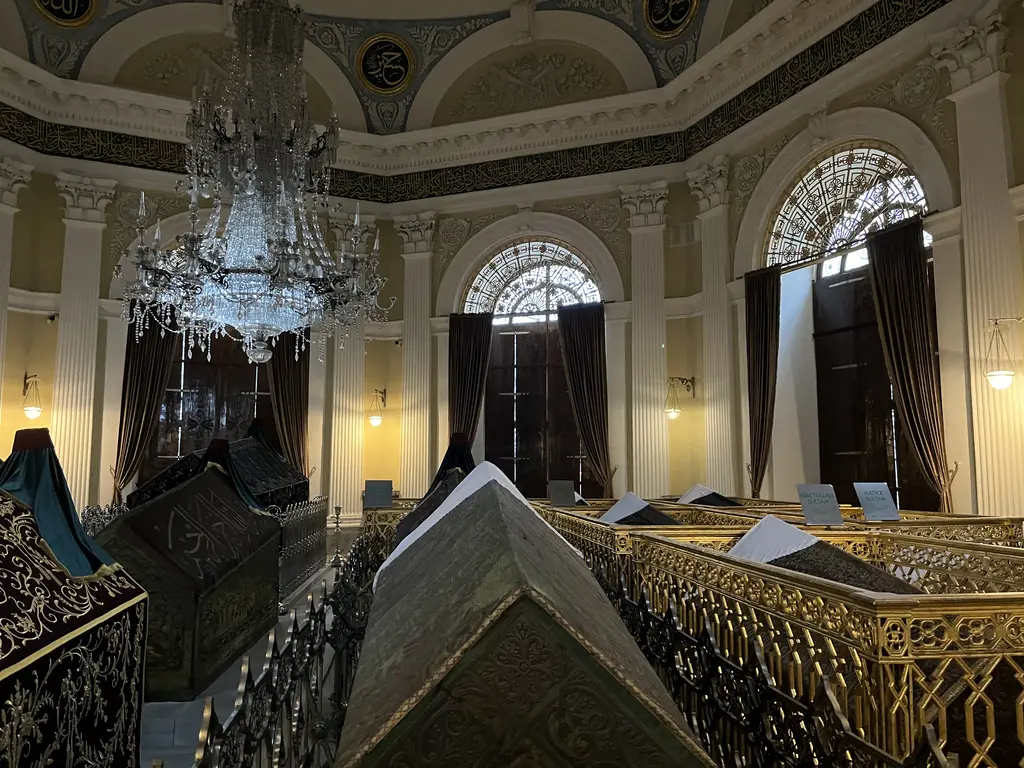 II. Mahmud Türbesi'nde kristal avize; altında ahşap parmaklıklar içinde yeşil ve siyah örtülü sandukalar, kemerli pencereler ve krem rengi duvarlar.