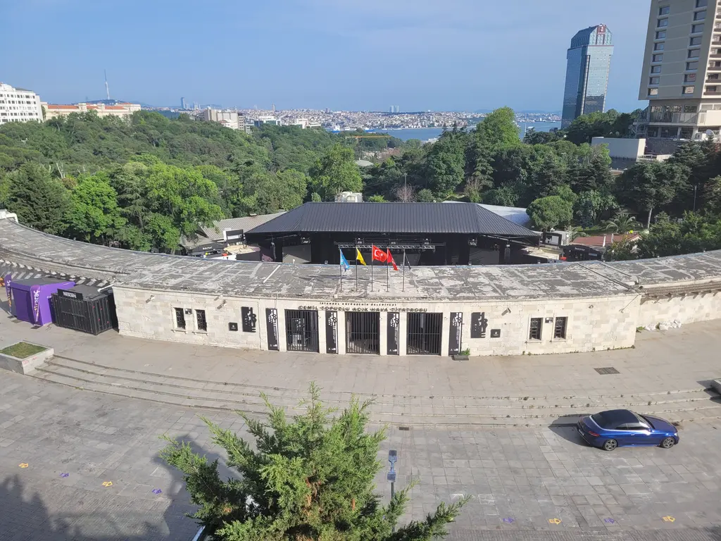 Harbiye'de Cemil Topuzlu Açıkhava Tiyatrosu; yarım daire taş basamaklar, sahne binası, ağaçlık yamaç ve uzakta Boğaz.