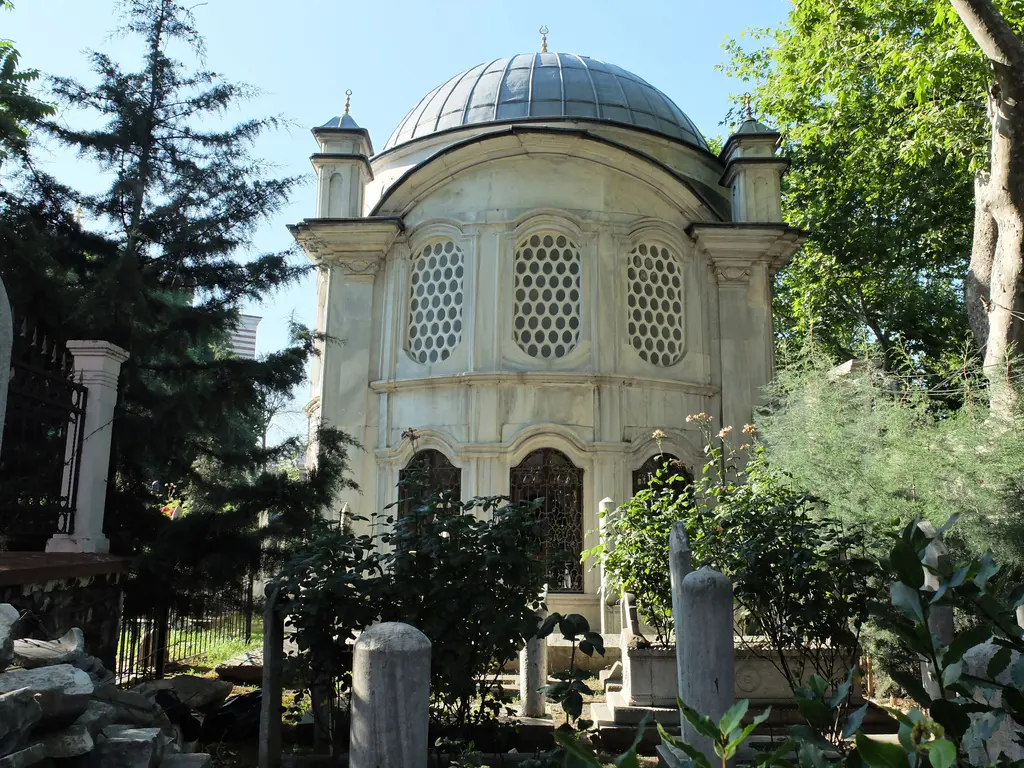 Şah Sultan Türbesi hazireden; yuvarlak kubbeli mermer yapı, petekli pencereler, ağaçlar ve önde eski mezar taşları.
