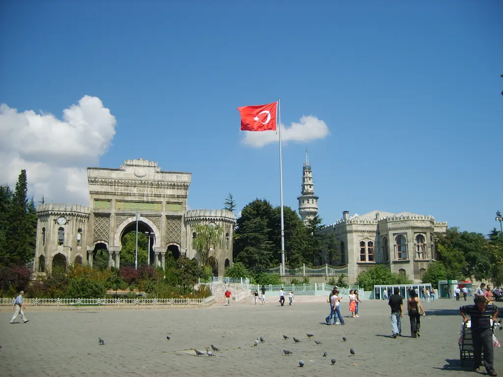 İstanbul Üniversitesi'nin taç kapısı ve Beyazıt Kulesi; kemerli anıtsal kapı, iki yanda Türk bayrakları ve arkasında yükselen kule.