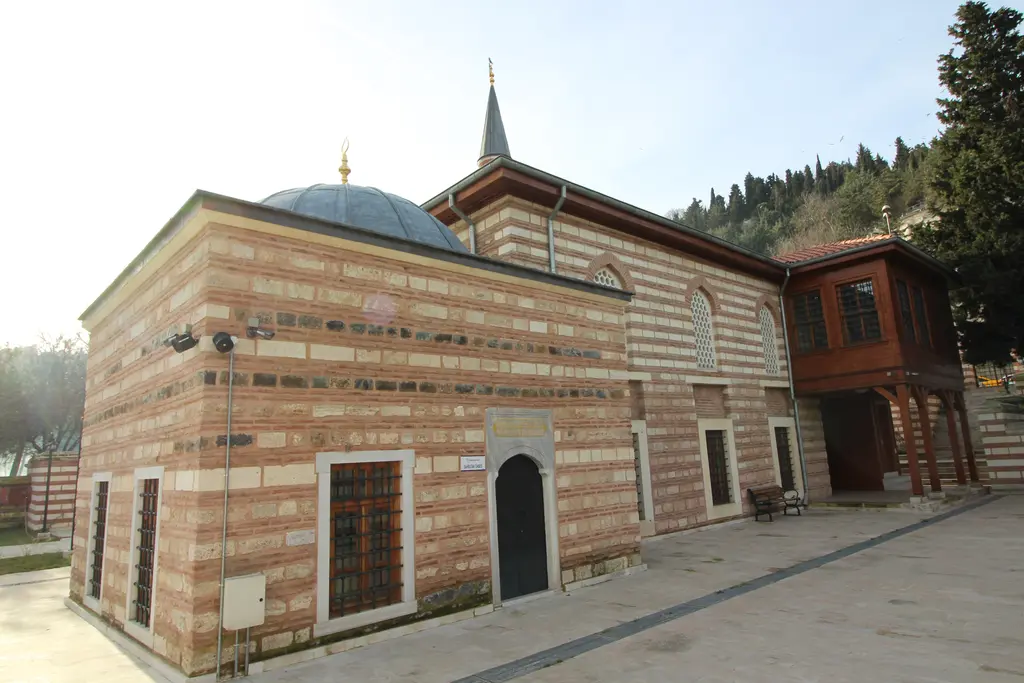 Şah Sultan Camii'nin dış cephesi; tuğla ve kesme taş sıralı duvarlar, kubbeli türbe, ahşap bölüm ve geniş taş avlu.