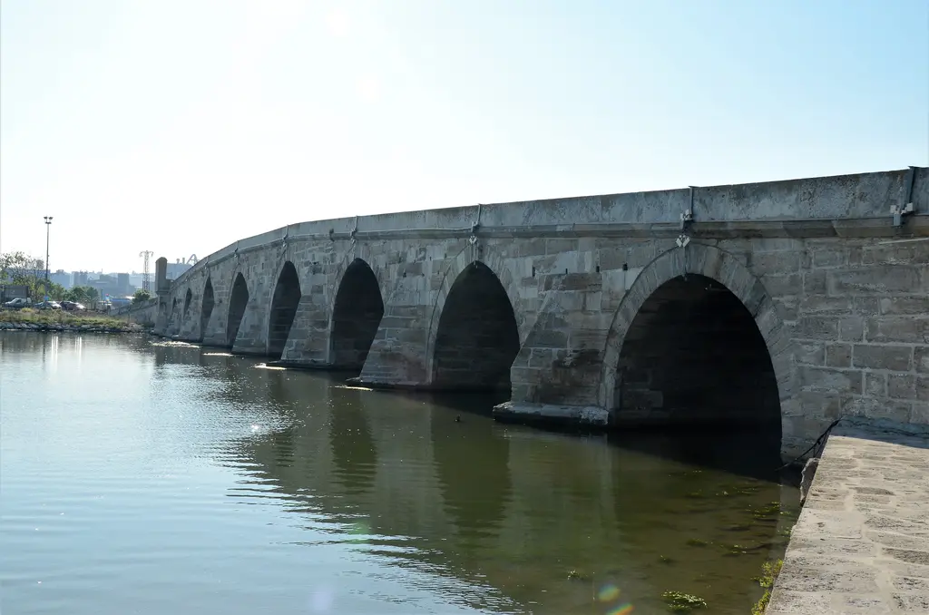 Kanuni Sultan Süleyman Köprüsü; büyüyen sivri kemerler, durgun suya yansımaları ve uzakta binalar.