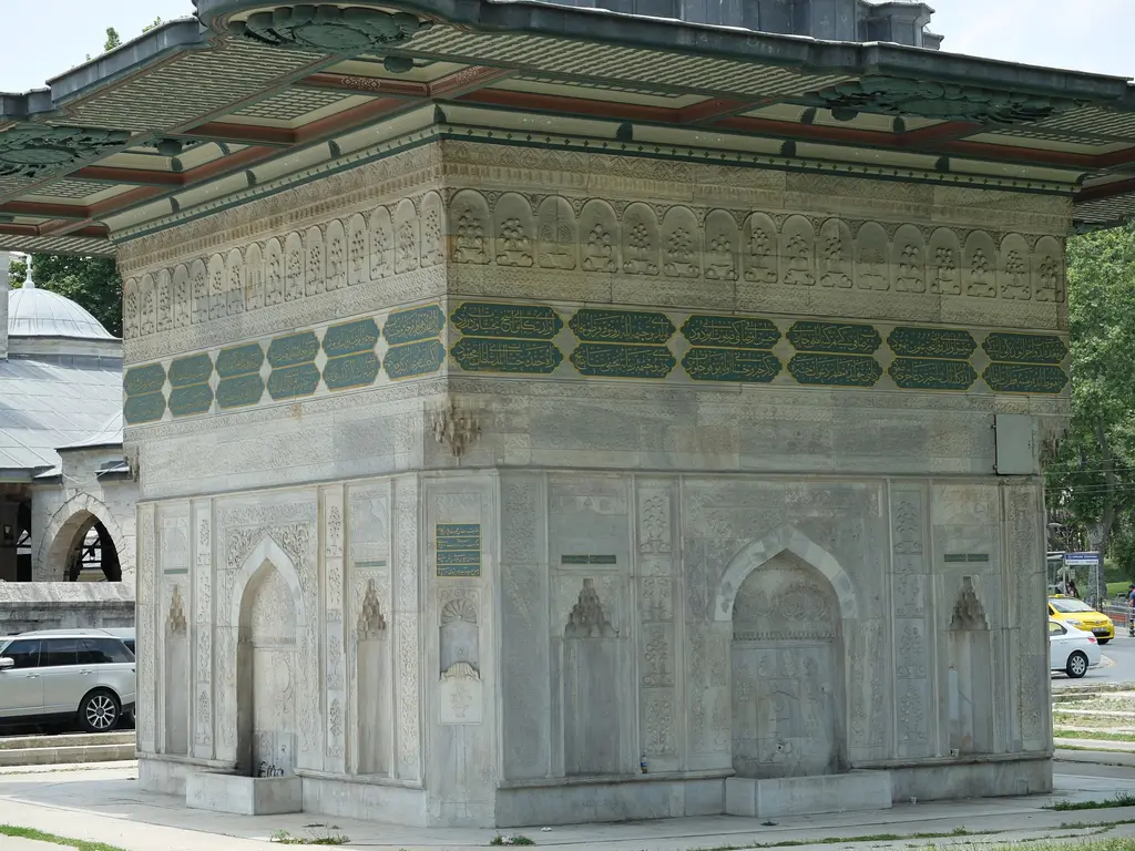 Tophane Çeşmesi'nin mermer gövdesi; nişler, yeşil kartuşlu yazı kuşağı, kabartmalı yüzeyler ve üstte saçağın gölgesi.
