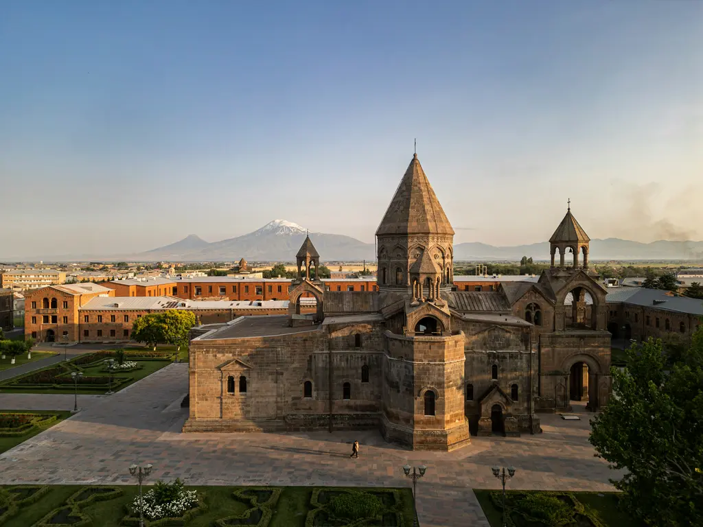Eçmiyadzin'de geniş bir avlunun ortasındaki taş katedral; konik külahlı merkezi kule, iki yanında çan kuleleri, çevresinde bahçeler ve arkada ovanın ardında Ağrı Dağı'nın karlı zirvesi.