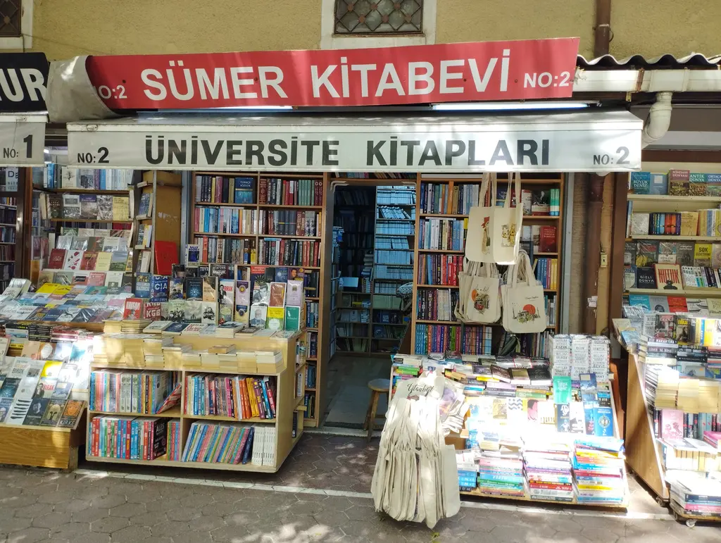 Sahaflar Çarşısı'nda kırmızı tabelalı bir kitabevi; altında beyaz "üniversite kitapları" bandı, kapının iki yanı tavana kadar raf, tavandan sarkan bez çantalar ve önde geniş kitap sergisi.