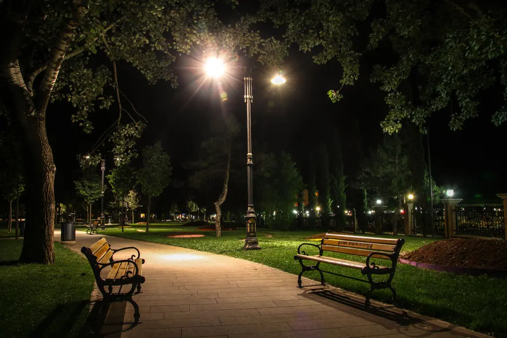 Göztepe Parkı'nda gece; aydınlatılmış fener direkleri, iki yanda ahşap banklar, ağaçlar ve taş kaplı yol.