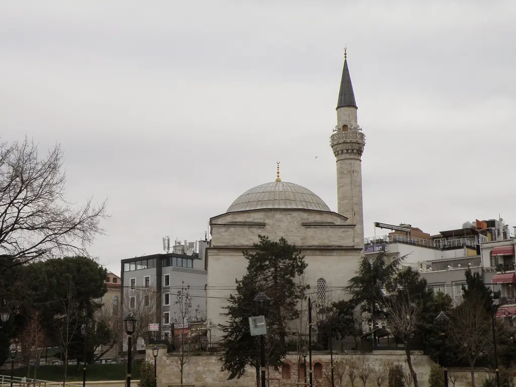 Bulutlu bir günde Firuz Ağa Camii; kesme taş gövde, kurşun kubbe, ince minare, önde ağaçlar ve yan tarafta binalar.