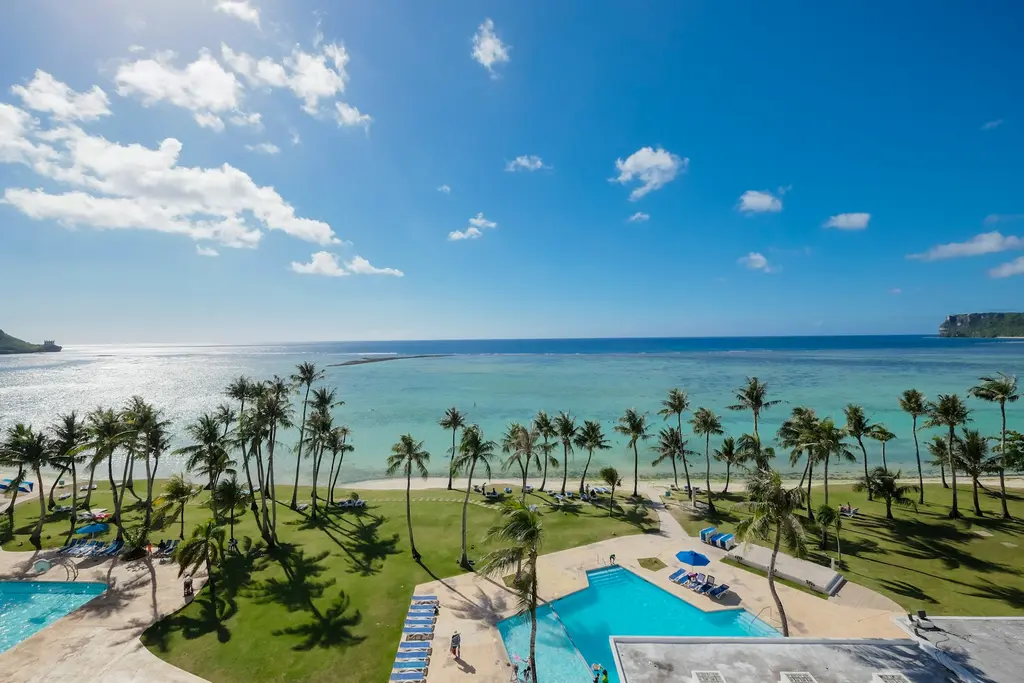 Guam'da Tumon Körfezi; palmiyeli çimenli sahil şeridi, otel havuzları ve sakin turkuaz koy, üstte parlak mavi gökyüzü ve dağınık bulutlar.