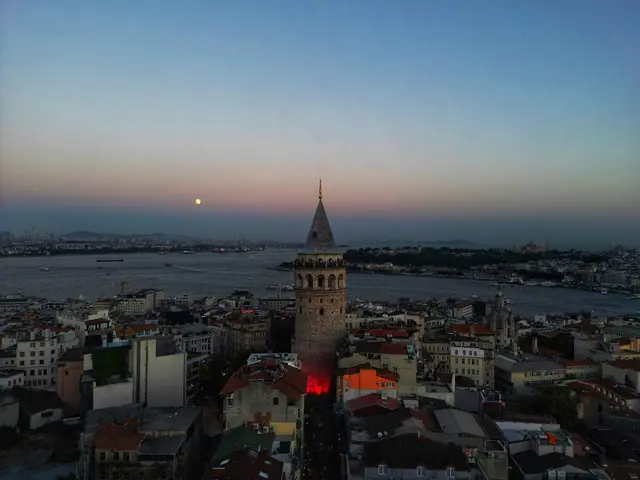 Havadan Galata Kulesi alacakaranlıkta; önde kiremitli çatılar ve kuleye uzanan dar sokak, arkada Haliç'in ağzı, tarihi yarımada ve ufukta dolunay.