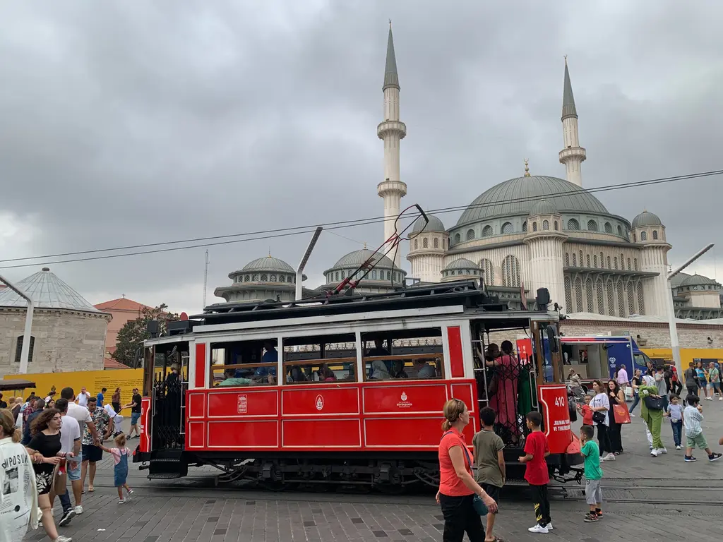 Taksim'de kırmızı nostaljik tramvay ve arkasında Taksim Camii; tramvayın yanında yolcular, geride kubbeler ve minareler.