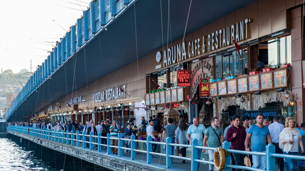 Galata Köprüsü'nün alt kat yürüyüş yolu; mavi korkuluk, kalabalık yayalar, lokanta tabelaları ve üstte köprü tabliyesi.
