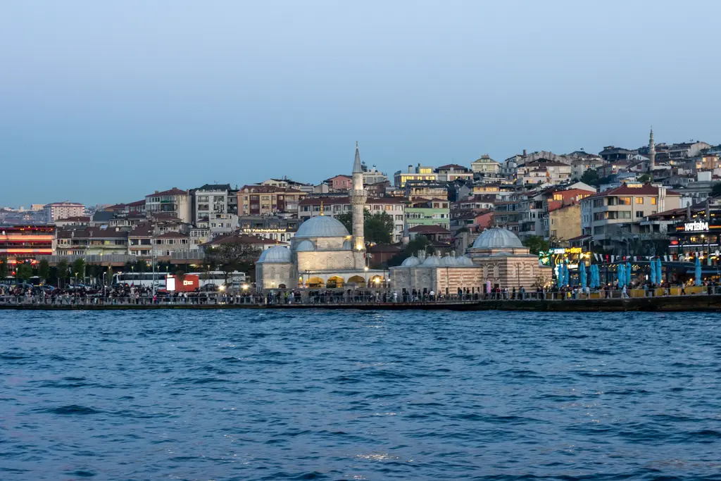 Denizden Üsküdar kıyısı; deniz kenarında kubbeli küçük cami ve medrese, arkasında yamaçlara tırmanan renkli evler.