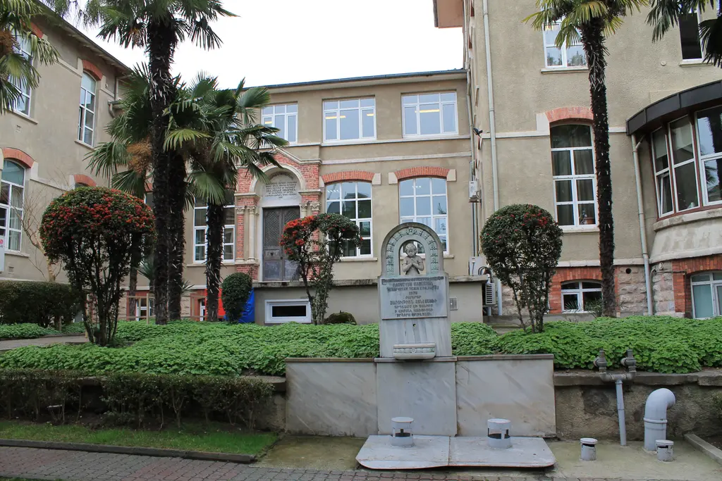 Surp Pırgiç Ermeni Hastanesi'nin bahçesi; tuğla ve taş cepheli tarihi bina, palmiyeler, budanmış çalılar ve mermer bir anıt.