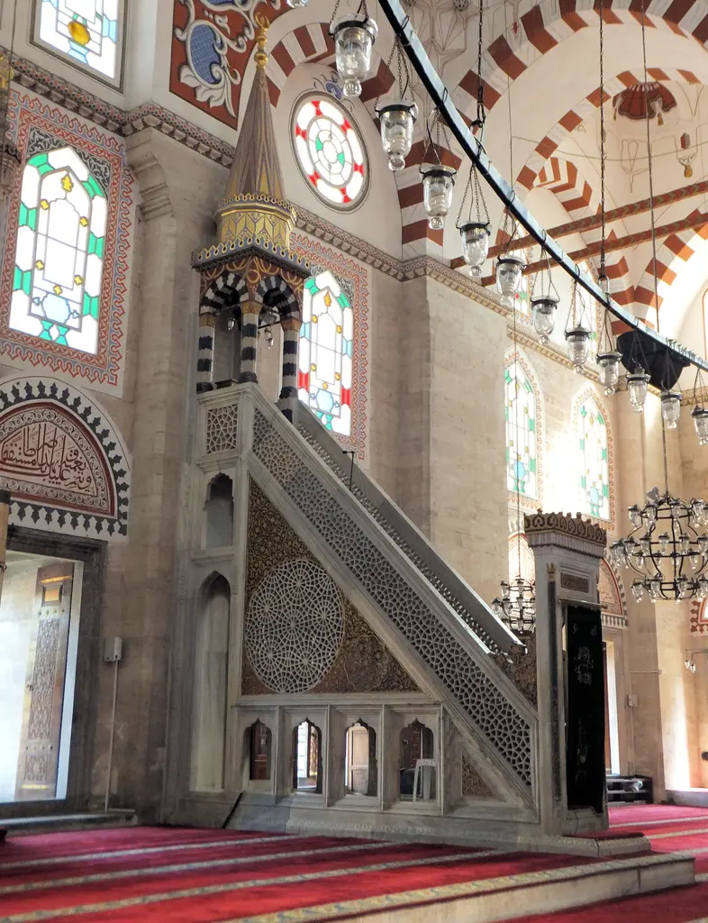 Şehzade Camii'nin minberi yakından; oyma şebekeli mermer korkuluk, külahlı köşk, asılı cam kandiller ve arkada renkli camlı pencere.