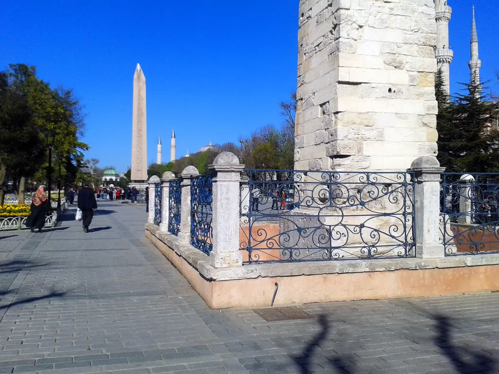 Örme Sütun'un parmaklıklı çevresi; koyu mavi gökyüzü, uzakta Dikilitaş ve minareler, taş döşeli yolda yürüyen insanlar.