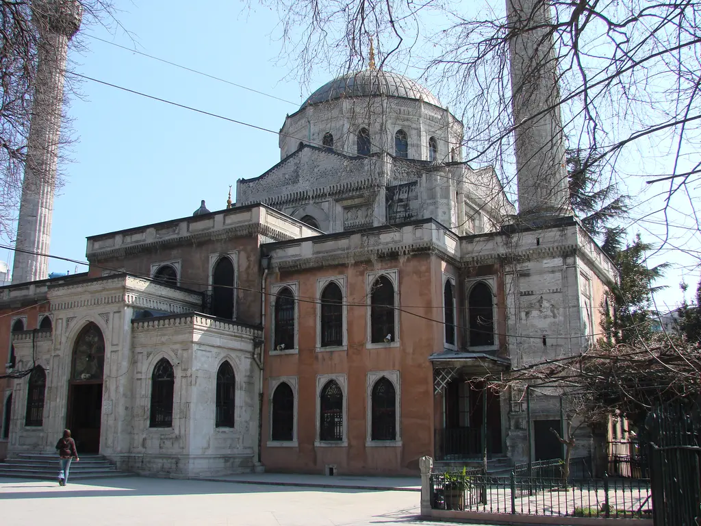 Pertevniyal Valide Sultan Camii ve yanındaki tuğla yapı; gotik pencereli taş cephe, minare, kubbe ve önünde taş döşeli avlu.