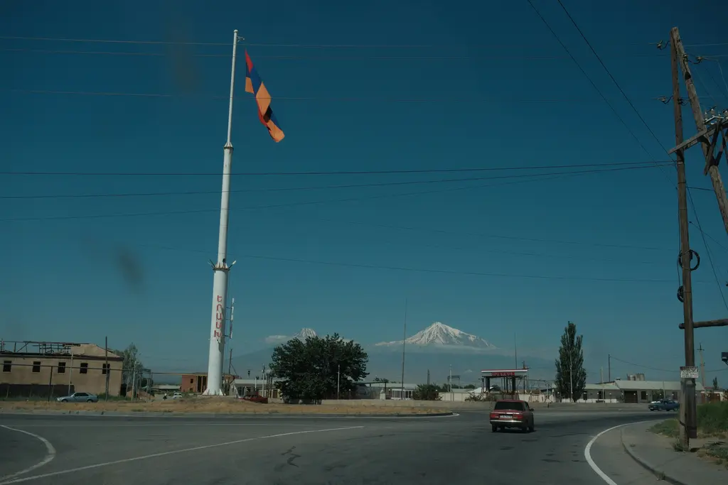 Ermenistan'da yol kenarındaki kavşakta dev bir direğe çekilmiş Ermenistan bayrağı; arkada ovanın ötesinde yükselen Ağrı Dağı'nın karlı zirvesi ile solunda Küçük Ağrı, önde asfalt yol ve alçak taş binalar.