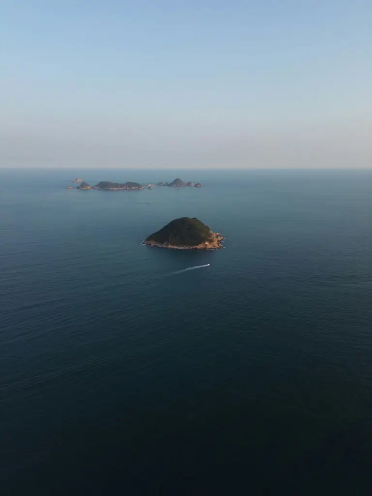 Sisli mavi bir denizde yükselen küçük yeşil adalar; ön plandaki adanın yanından iz bırakarak geçen küçük bir tekne, arkada bir takımada.