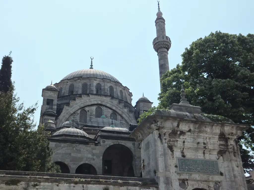 Ayazma Camii'nin avlusundan görünüm; önde taş kapı yapısı ve ahşap kapı, arkada kubbe, minare ve ağaç.
