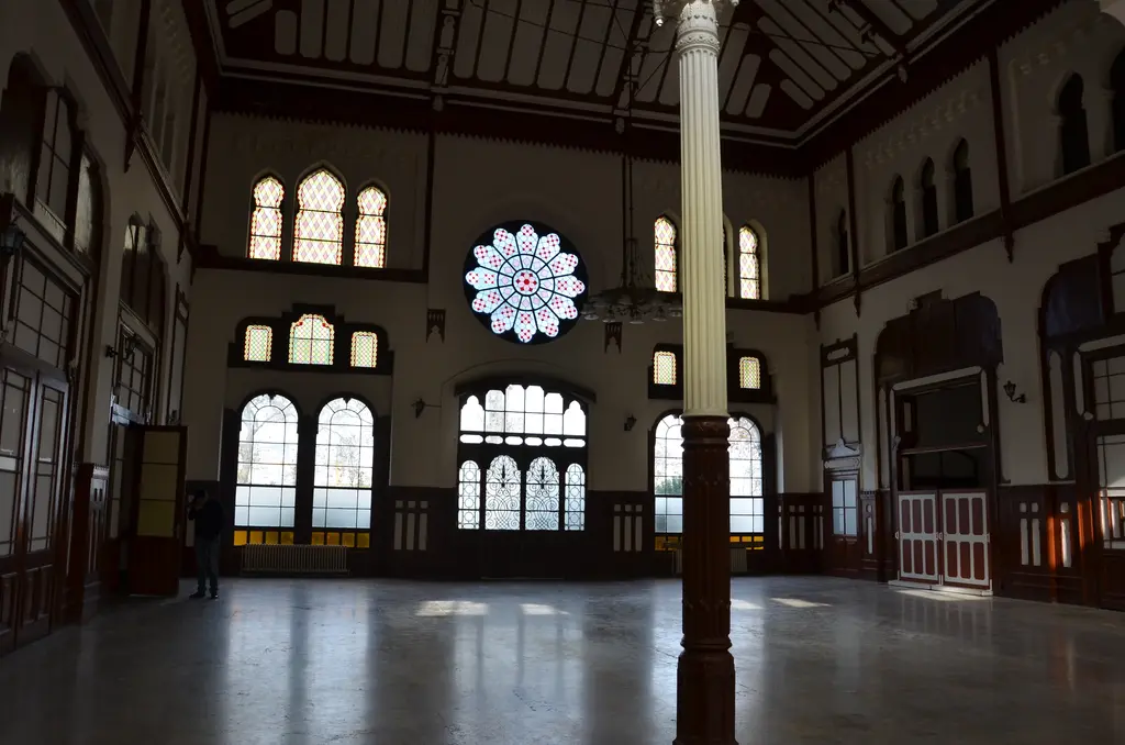 Sirkeci Garı'nın büyük salonu boşken; koyu parlak zemin, arka duvarda yüksekte mavi beyaz gül pencere, altında yuvarlak kemerli uzun pencere dizisi ve ahşap kaplama.