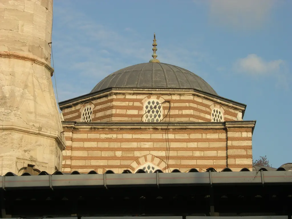 Koca Mustafa Paşa Camii'nin sekizgen kubbe kasnağı akşam ışığında; dönüşümlü taş ve tuğla bantlar, kemerli şebekeli pencereler, gri kurşun kubbe, altın alem ve solda minare gövdesi.