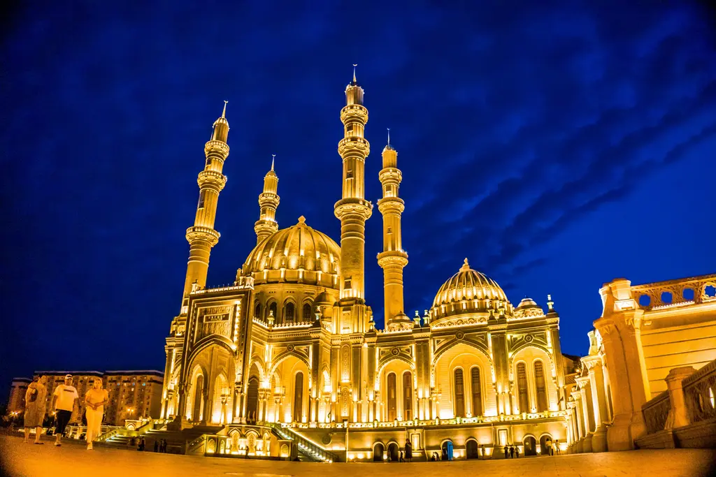 Bakü'de Heydər Camii'nin gece görünümü; altın sarısı aydınlatmayla parlayan kubbeler, dört ince minare ve merdivenli geniş giriş cephesi.