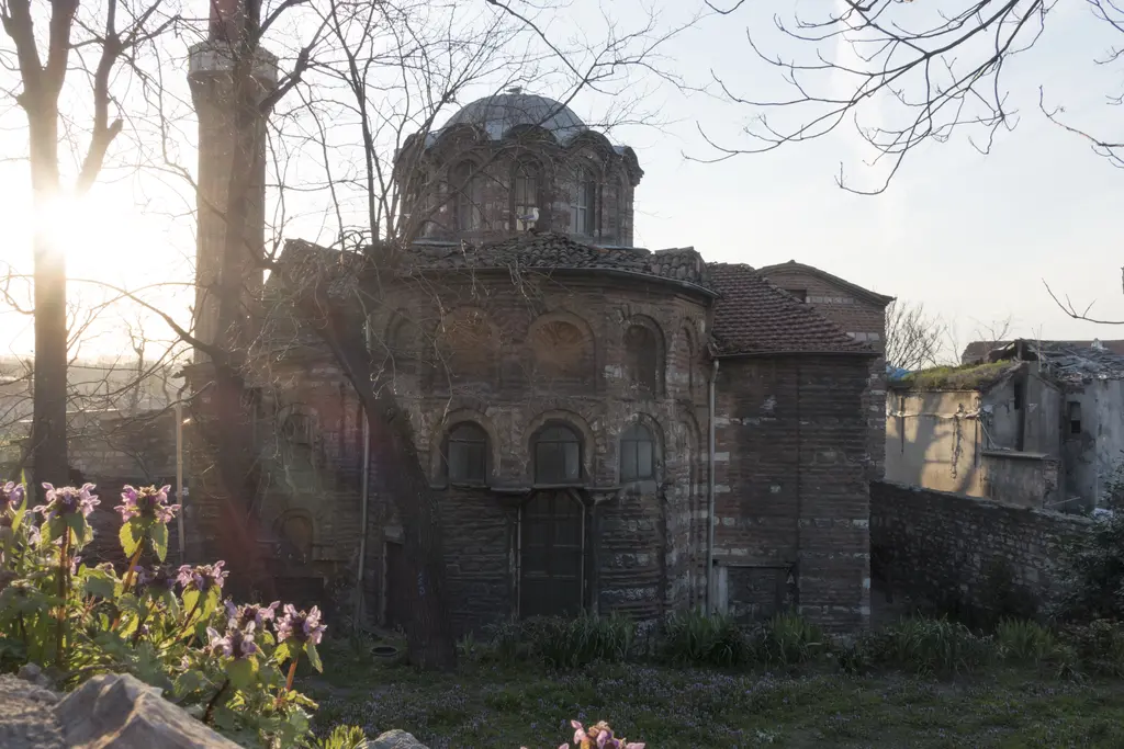 Vefa Kilise Camii doğudaki boş bahçeden gün batımında; ağaçların arasından gelen ışıkta silüetleşen kütle, kubbe kasnağı, yuvarlak doğu ucu ve uzun kemerli pencereler.
