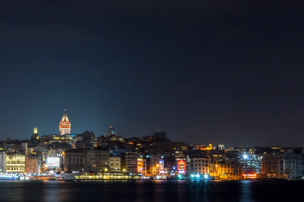 Gece Karaköy ve Galata yamacı; ışıklı Galata Kulesi, kıyıdaki aydınlatılmış binalar ve suya yansıyan ışıklar.