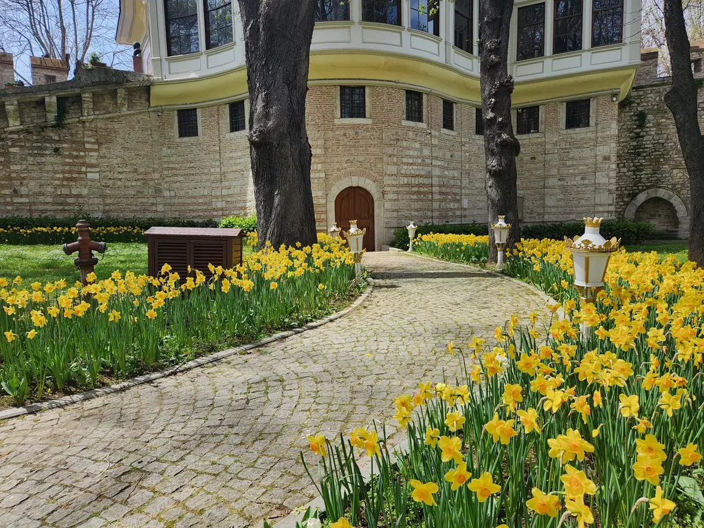 Gülhane Parkı'nda Alay Köşkü'nün surun üstündeki kanadı; önde sarı nergis tarhları, parke taş yol, fenerler ve ağaç gövdeleri.