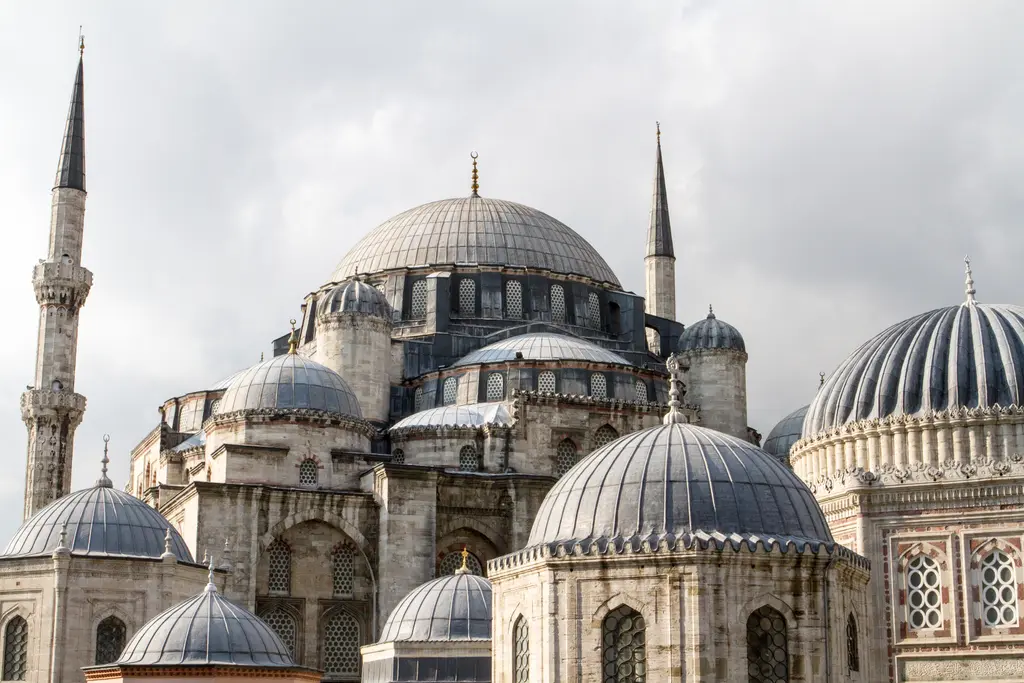 Şehzade Camii'nin kubbe kümesi yakından; merkezî kubbe ile çevresindeki yarım kubbeler, solda minare gövdesi, sağda dilimli türbe kubbesi, gri bulutlu gökyüzü.